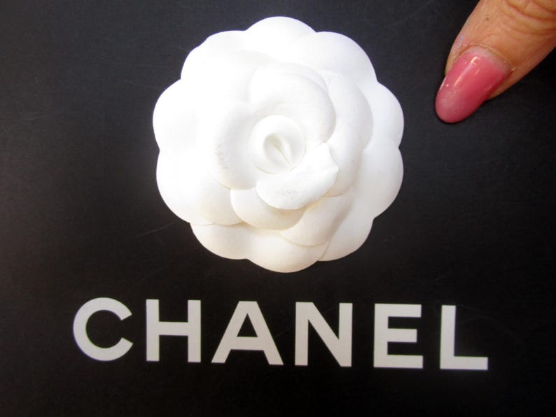 May include: A white camellia flower on a black surface with the word "CHANEL" in white block letters. The flower has multiple layers of petals. A pink fingernail is visible in the image.
