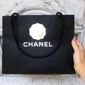 May include: Black shopping bag with a white camellia flower and the word "CHANEL" in white lettering. The bag has a black rope handle. The bag is held by a person.