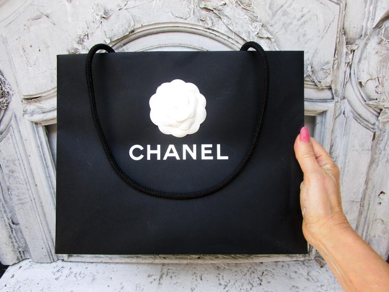 May include: Black shopping bag with a white camellia flower and the word "CHANEL" in white lettering. The bag has a black rope handle. The bag is held by a person.