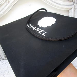 May include: Black shopping bag with the word "CHANEL" in white, along with a white logo. The bag has a black rope handle. The bag is held by a person.