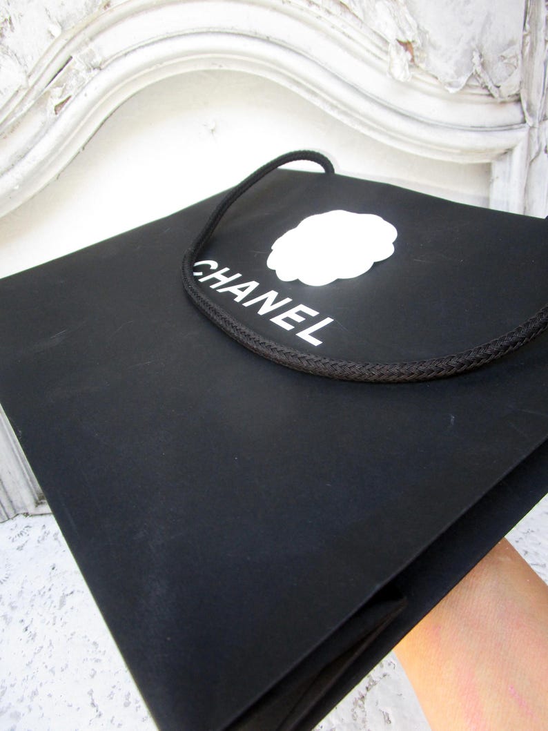 May include: Black shopping bag with the word "CHANEL" in white, along with a white logo. The bag has a black rope handle. The bag is held by a person.