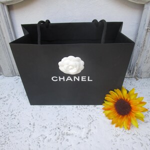 May include: Black shopping bag with the word "CHANEL" in white, a white flower emblem, and black rope handles. A yellow and orange sunflower rests beside the bag.