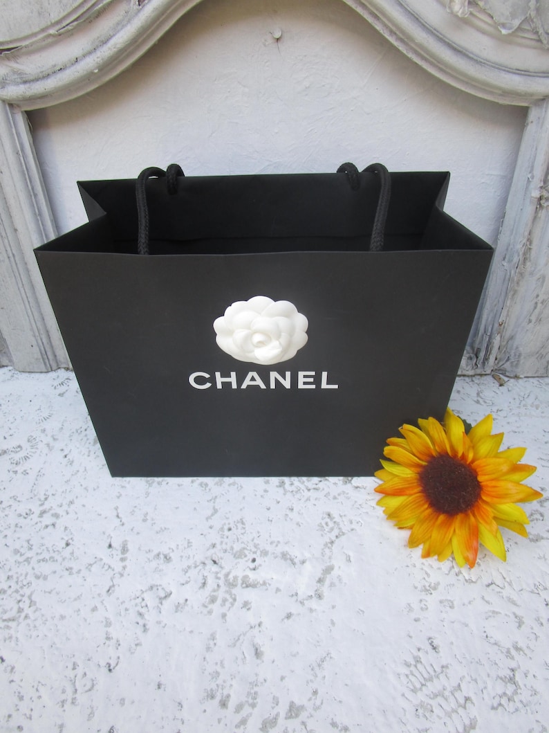 May include: Black shopping bag with the word "CHANEL" in white, a white flower emblem, and black rope handles. A yellow and orange sunflower rests beside the bag.