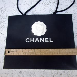 May include: Black shopping bag with white "CHANEL" text and a white flower emblem. The bag has black rope handles and a wooden ruler is placed across the bottom. The bag is approximately 13 inches wide.