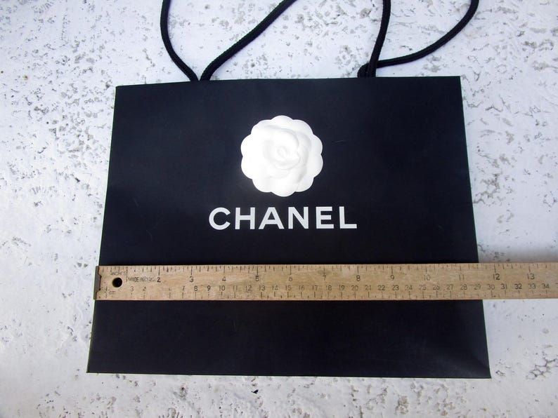 May include: Black shopping bag with white "CHANEL" text and a white flower emblem. The bag has black rope handles and a wooden ruler is placed across the bottom. The bag is approximately 13 inches wide.