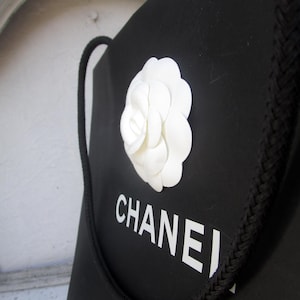 May include: Black shopping bag with a white camellia flower and the word "CHANEL" in white block letters. The bag has a black rope handle and a braided trim along the top edge.