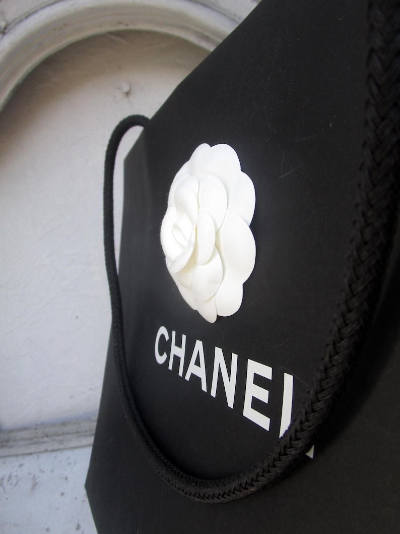 May include: Black shopping bag with a white camellia flower and the word "CHANEL" in white block letters. The bag has a black rope handle and a braided trim along the top edge.