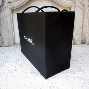 May include: A black shopping bag with the word "CHANEL" printed in white. The bag has black rope handles and is sitting on a white surface.