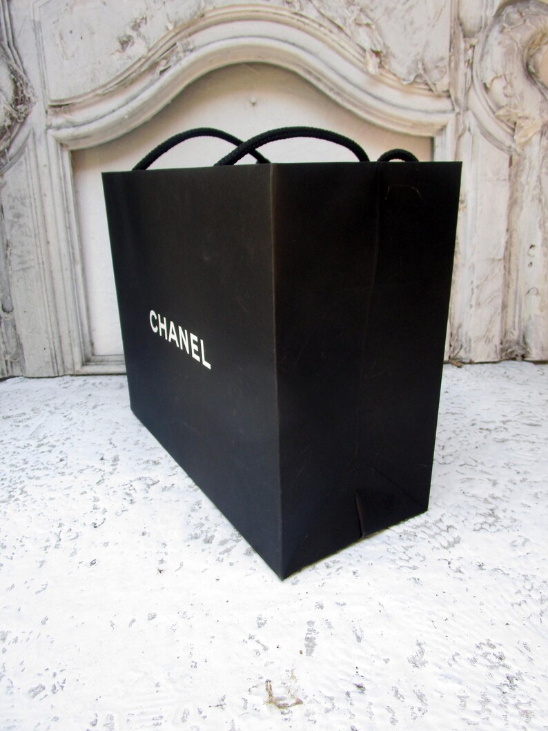 May include: A black shopping bag with the word "CHANEL" printed in white. The bag has black rope handles and is sitting on a white surface.