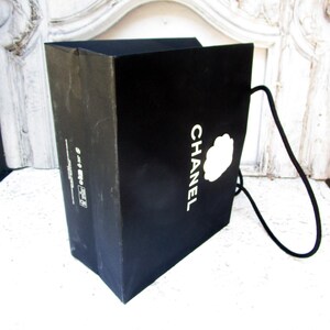 May include: A black shopping bag with the word "CHANEL" in white lettering and a white camellia flower logo. The bag has a black cord handle and is made of a sturdy material.