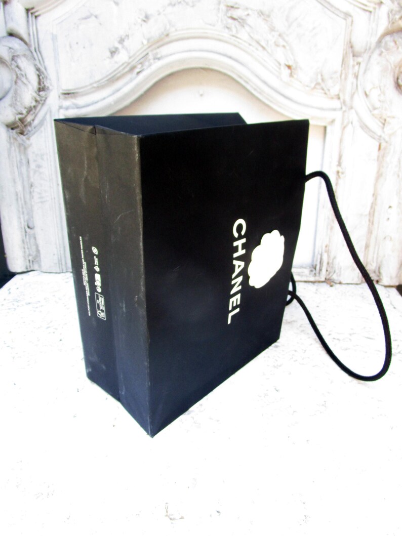 May include: A black shopping bag with the word "CHANEL" in white lettering and a white camellia flower logo. The bag has a black cord handle and is made of a sturdy material.