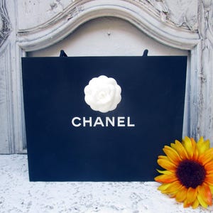 May include: A navy blue shopping bag with the word "CHANEL" in white lettering and a white camellia flower. A yellow and orange sunflower is placed to the right of the bag.