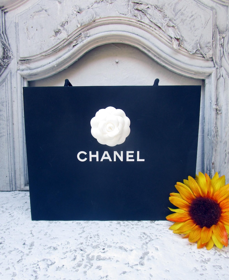 May include: A navy blue shopping bag with the word "CHANEL" in white lettering and a white camellia flower. A yellow and orange sunflower is placed to the right of the bag.