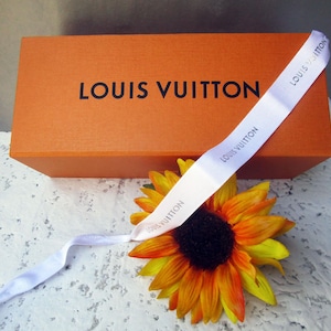 May include: An orange Louis Vuitton box with the brand name in black lettering. A white ribbon with the brand name is draped across the box. A yellow and orange sunflower is in the foreground.