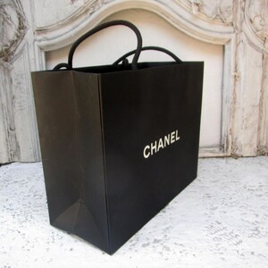 May include: A black shopping bag with the word "CHANEL" printed in white. The bag has black rope handles and is sitting on a white surface.
