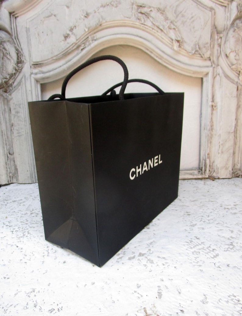 May include: A black shopping bag with the word "CHANEL" printed in white. The bag has black rope handles and is sitting on a white surface.