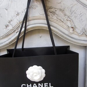 May include: Black shopping bag with a white camellia flower emblem and the brand name "CHANEL" in white lettering. The bag has black rope handles and is held up by a person.