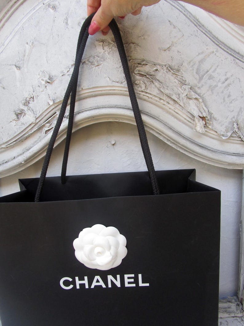 May include: Black shopping bag with a white camellia flower emblem and the brand name "CHANEL" in white lettering. The bag has black rope handles and is held up by a person.