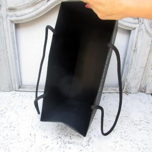 May include: A black gift bag with black rope handles. The bag is held open, revealing the interior. The background is a white surface and a white and gray decorative frame.