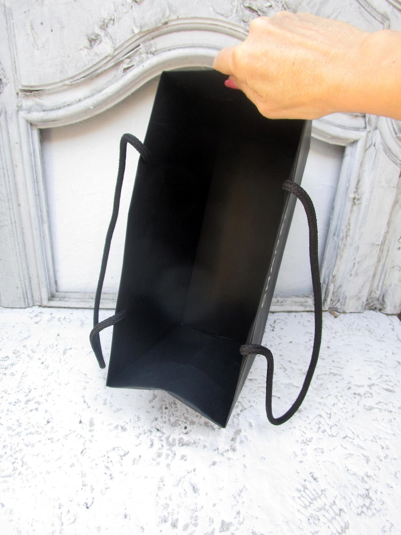 May include: A black gift bag with black rope handles. The bag is held open, revealing the interior. The background is a white surface and a white and gray decorative frame.