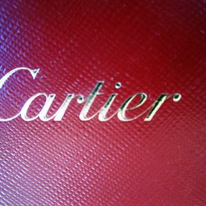 Może przedstawiać: Ciemnoczerwona, teksturowana powierzchnia z wytłoczonym napisem Cartier w metalicznym złotym kroju pisma. Materiał posiada wz&oacute;r w kratkę, kt&oacute;ry tworzy subtelny efekt siatki na całym tle, podkreślając nazwę marki.