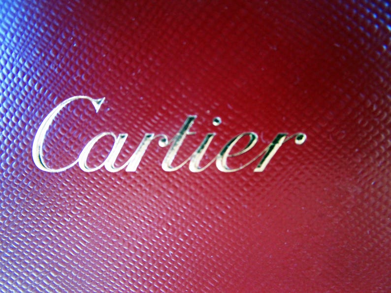 Może przedstawiać: Ciemnoczerwona, teksturowana powierzchnia z wytłoczonym napisem Cartier w metalicznym złotym kroju pisma. Materiał posiada wz&oacute;r w kratkę, kt&oacute;ry tworzy subtelny efekt siatki na całym tle, podkreślając nazwę marki.