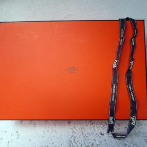 May include: An orange rectangular box with a black border and a dark ribbon. The ribbon has white text that reads "HERMES - PARIS". The box has a small logo in the center.