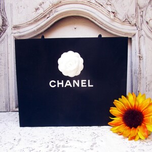 May include: A black shopping bag with the word "CHANEL" in white lettering. A white camellia flower is centered above the brand name. A yellow and orange sunflower is to the right of the bag.