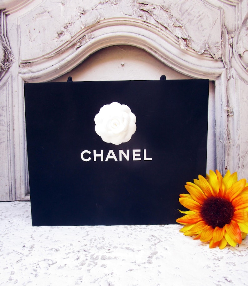 May include: A black shopping bag with the word "CHANEL" in white lettering. A white camellia flower is centered above the brand name. A yellow and orange sunflower is to the right of the bag.