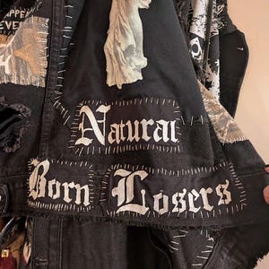 Op de afbeelding: Zwarte spijkerjas met patches en witte stiksels. Patches met de woorden "Natural Born Losers" in gotische letters en een witte afbeelding van een figuur. De jas heeft een versleten uitstraling.