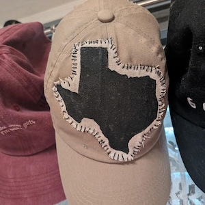 Puede incluir: Una gorra de béisbol de color tostado con un parche de tela negro en forma del estado de Texas. El parche está sujeto a la parte delantera de la gorra con puntadas de festón visibles, gruesas y oscuras alrededor de todo el contorno.
