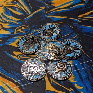 May include: A collection of silver-colored, round coins with intricate designs and light blue accents. The coins feature various stylized images and symbols. They are arranged on a patterned surface with blue and yellow hues.