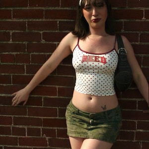 May include: A white spaghetti strap tank top with red polka dots and the word REED printed in red block letters across the chest, paired with a dark olive green camouflage print denim mini skirt and a black shoulder bag with a silver buckle.