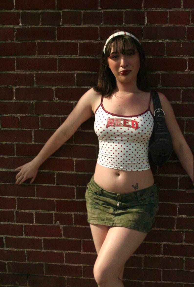 May include: A white spaghetti strap tank top with red polka dots and the word REED printed in red block letters across the chest, paired with a dark olive green camouflage print denim mini skirt and a black shoulder bag with a silver buckle.