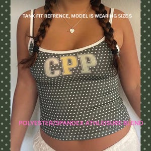 May include: A dark olive green tank top with a white polka dot pattern and white trim. The front features the letters CPP in a light-colored block font. Text on the image reads: TANK FIT REFRENCE, MODEL IS WEARING SIZE S and POLYESTER/SPANDEX ATHLEISURE BLEND.
