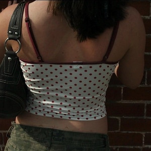 May include: A white camisole featuring a red polka dot pattern with thin maroon straps and trim, paired with a black shoulder bag that has a silver metal ring connector and visible white stitching along the dark fabric seams.