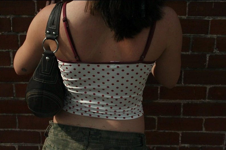 May include: A white camisole featuring a red polka dot pattern with thin maroon straps and trim, paired with a black shoulder bag that has a silver metal ring connector and visible white stitching along the dark fabric seams.