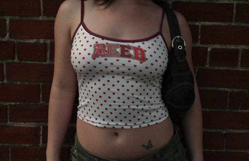 May include: A white spaghetti strap tank top featuring a red polka dot pattern and the word REED printed in red block letters across the chest. The garment has thin red straps and a red trim along the neckline, paired with a black shoulder bag.