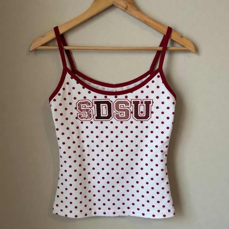May include: A white camisole with a repeating pattern of small maroon polka dots and thin maroon spaghetti straps. The front features the letters SDSU in a block font with a maroon outline and a textured, patterned fill, centered on the chest.