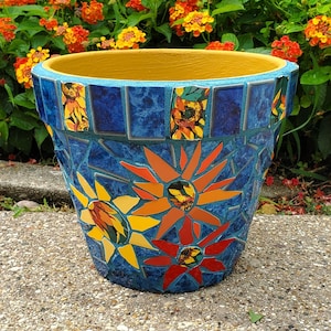 May include: A tapered flower pot featuring a mosaic design of blue tiles and floral patterns in yellow, orange, and red. The interior rim is painted a solid yellow, and the exterior surface is covered in a decorative arrangement of glass pieces.