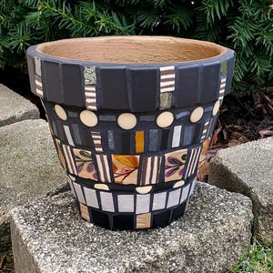 May include: A decorative mosaic flower pot with a black exterior, accented with beige, white, and patterned tiles. The pot features a brown interior and is sitting on a stone surface. The mosaic design includes rectangular and circular elements.