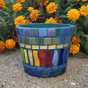 May include: A mosaic-tiled flower pot with a teal interior and exterior. The pot features a band of gold tiles and a rainbow of colors. The background includes vibrant orange flowers and green foliage.