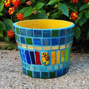 May include: A cylindrical planter featuring a vibrant mosaic exterior composed of square and rectangular glass tiles in shades of blue, green, yellow, and red, with a central floral accent piece and a bright yellow interior rim and basin.
