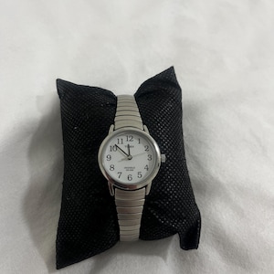 Könnte beinhalten: Eine silberfarbene Timex Indiglo-Uhr mit weißem Zifferblatt und schwarzen Ziffern. Die Uhr hat ein Stretchband und wird auf einem schwarzen Kissen präsentiert. Das Zifferblatt zeigt die Uhrzeit 10:09 Uhr an.