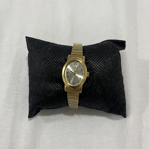 May include: A gold wristwatch with a rectangular face and a gold strap. The watch face displays the numbers 1 to 12 and the word "TIME". The watch is resting on a black cushion.