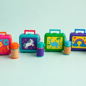 May include: Four miniature plastic lunch boxes with matching thermos containers. The first is pink with flowers, the second teal with a unicorn, the third green with a dinosaur, and the fourth purple with a rainbow. Each set features a handle.
