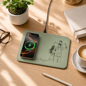 May include: A sage green rectangular wireless charging pad featuring a black line-art illustration of a family and the text "We love you!" printed on the surface. A smartphone is placed on the left side of the pad, displaying a green charging icon.