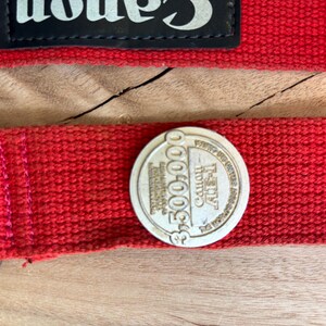 Puede incluir: Primer plano de una correa de cámara roja con el logotipo de Canon y una moneda conmemorativa. La moneda es circular y presenta el texto "$3.300.000" y "Canon AE-1". La correa es de material tejido.