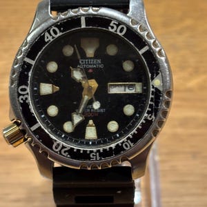 May include: A Citizen Automatic diver watch with a black dial, luminous hour markers, and a black rotating bezel with silver numerals. The watch features a day-date window at the three o'clock position, a gold-tone crown, and a black rubber strap.