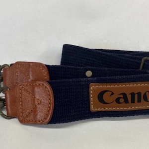 Puede incluir: Correa de cámara azul marino con detalles de cuero marrón y el nombre de la marca "Canon" en letras marrones. La correa tiene cierres metálicos y hebillas ajustables para una fijación segura.